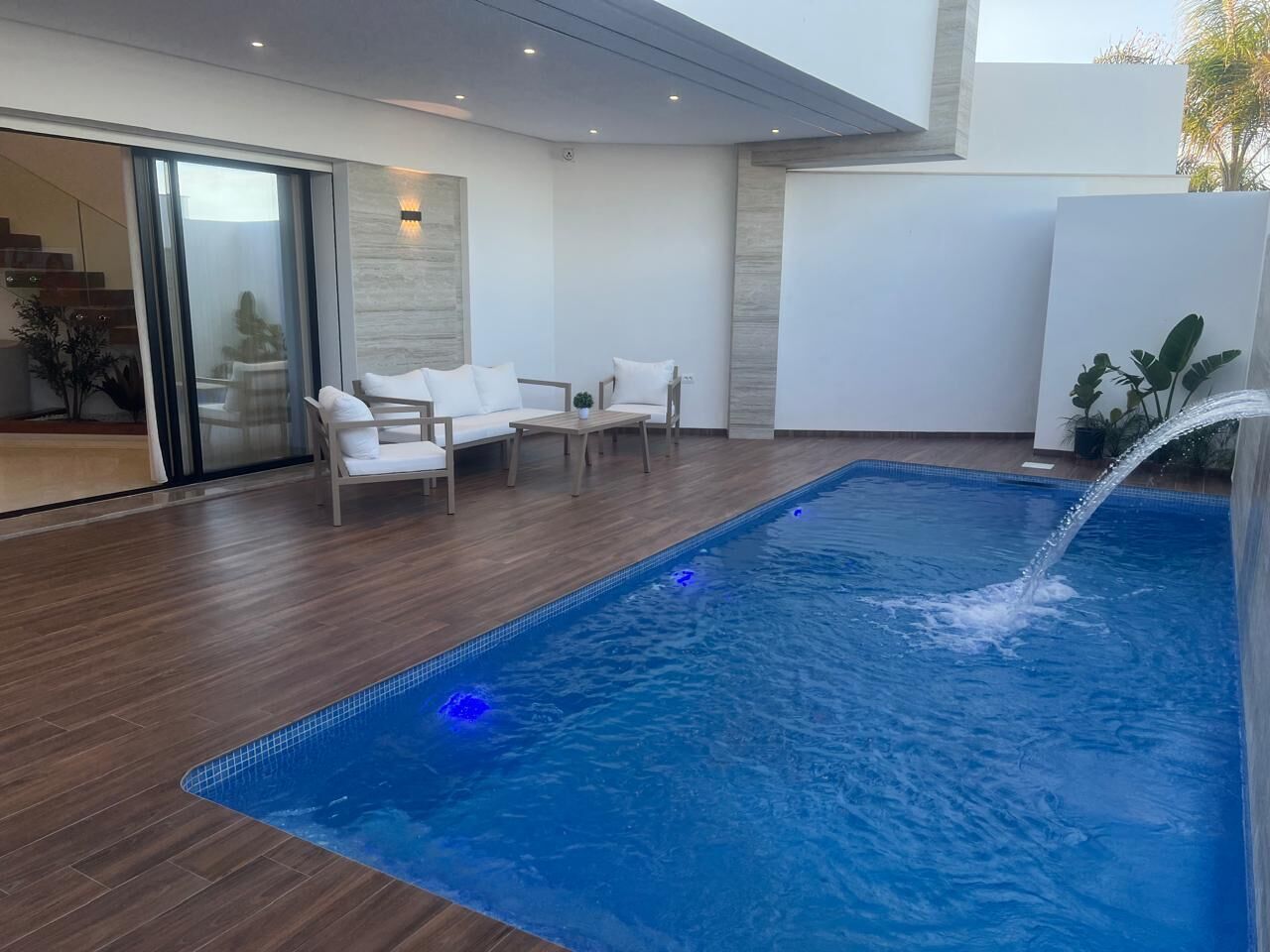 Una piscina con fuente acuática, rodeada por una terraza de madera y mobiliario blanco.