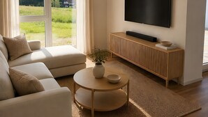 Een moderne woonkamer met een bank, een salontafel, een flatscreen televisie aan de muur en uitzicht op een groen landschap door grote ramen.