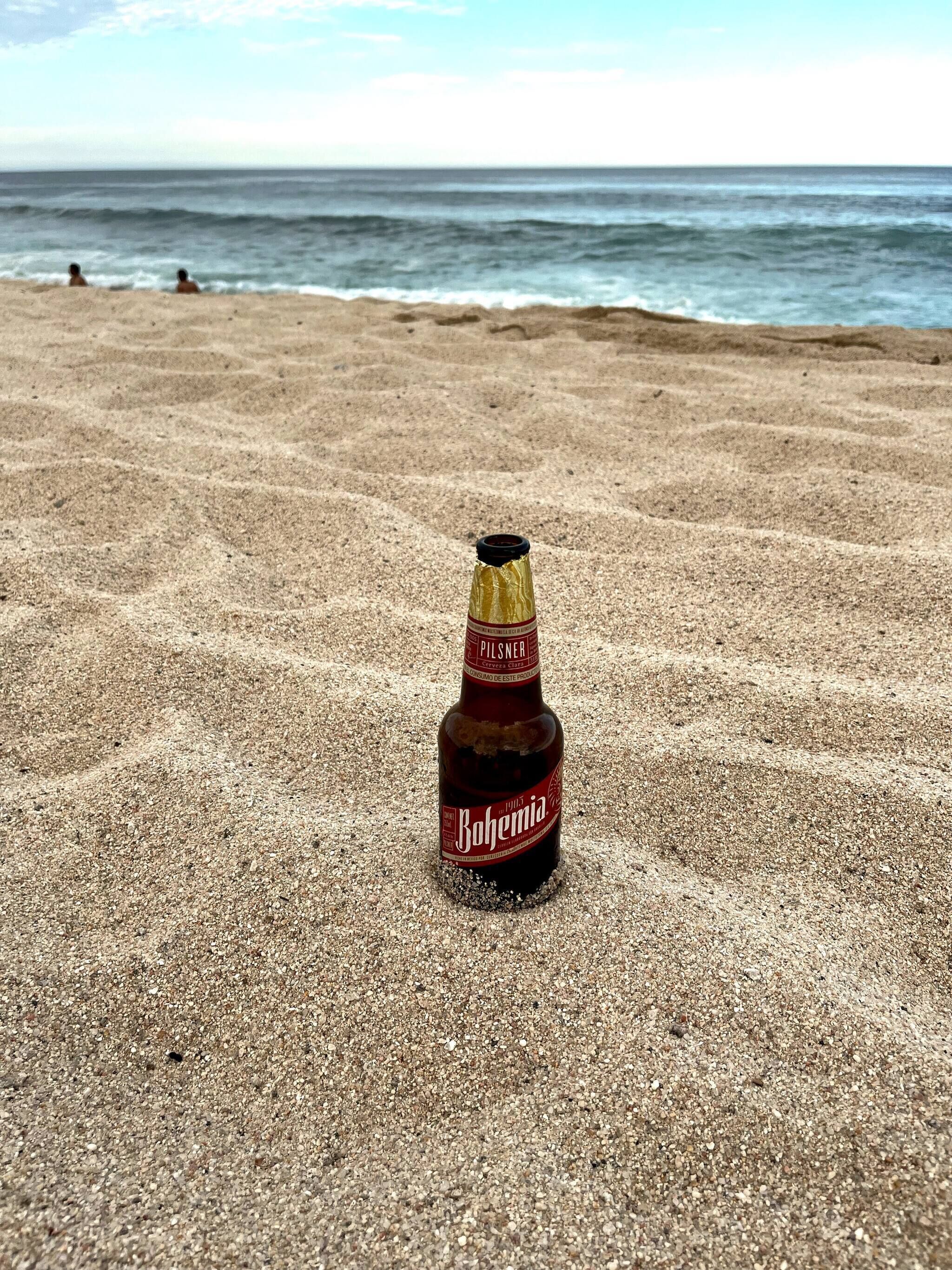 A bottle of Bohemia beer on a sandy beach with the ocean in the background.