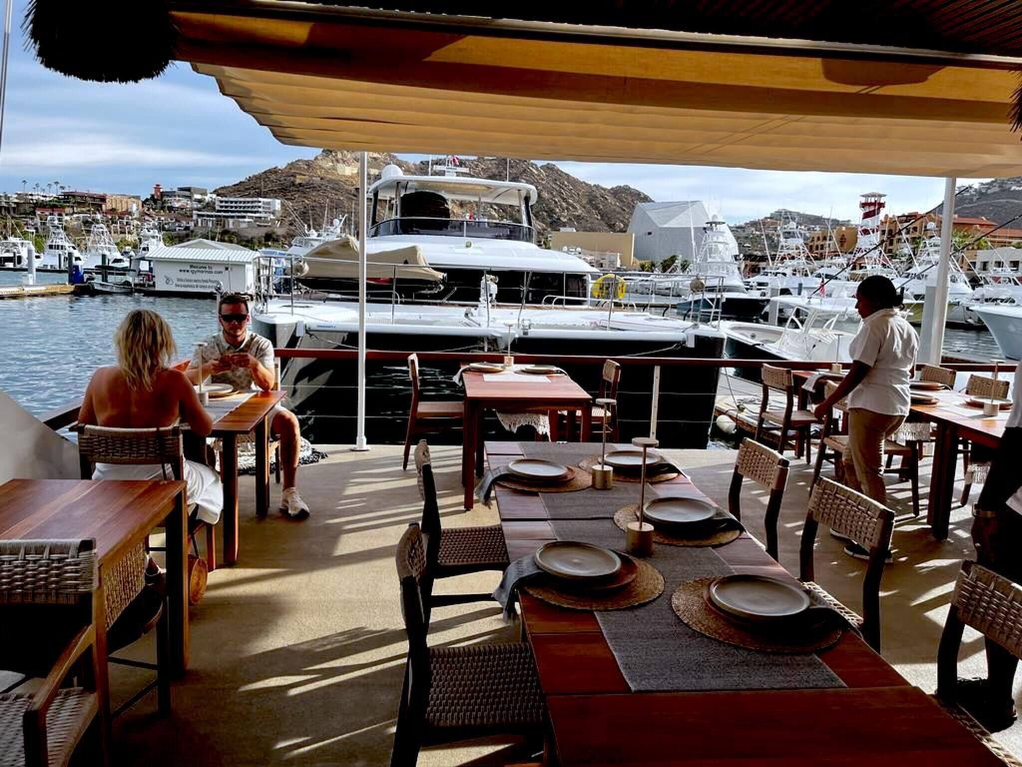 An outdoor dining area with tables and chairs, overlooking a marina with boats and a mountainous backdrop.