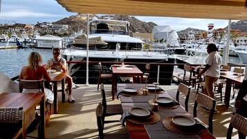 Un área de comedor al aire libre con mesas y sillas, con vista a un puerto deportivo con barcos y montañas al fondo.