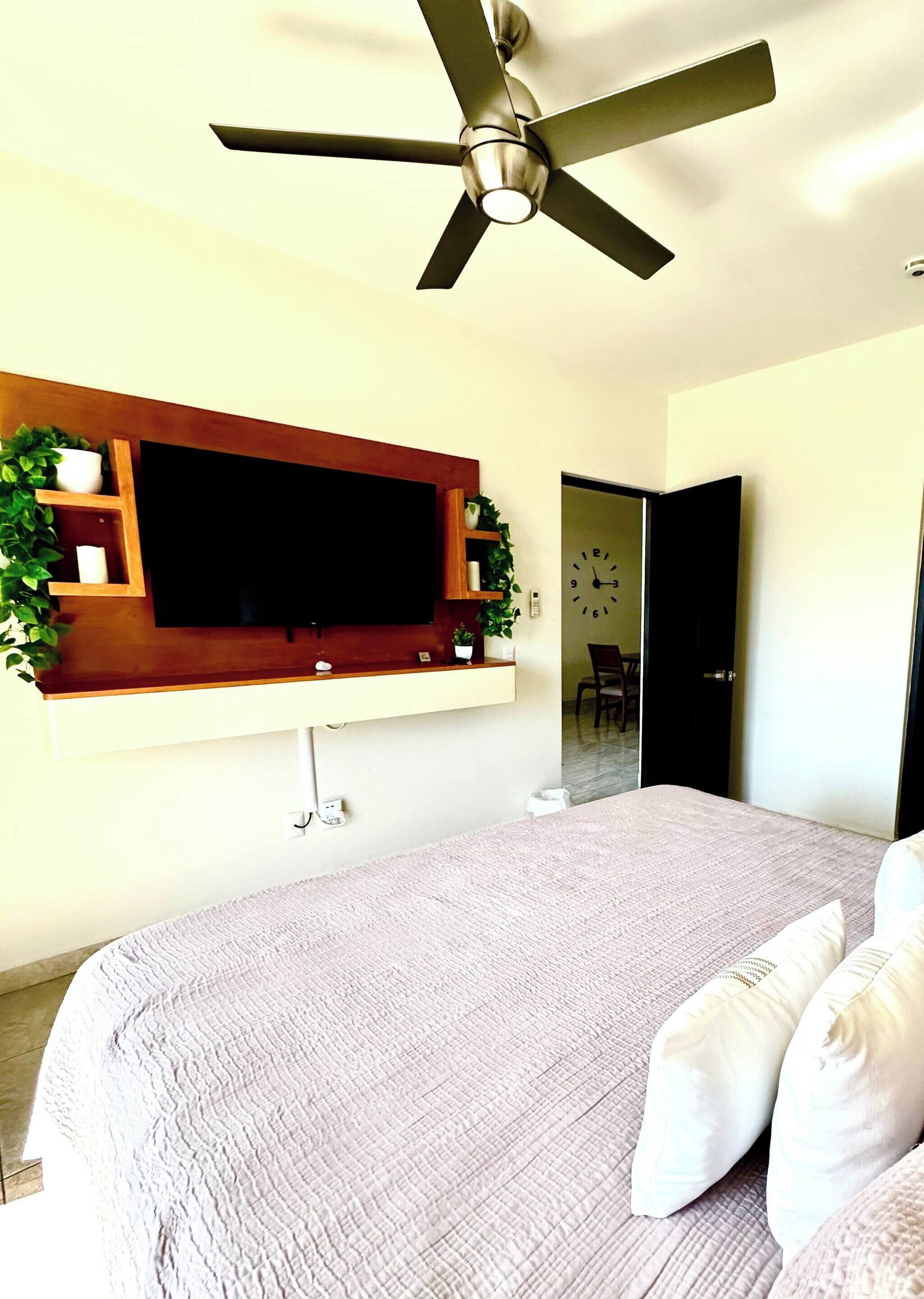 Un dormitorio con cama, ventilador de techo, televisor montado y puerta que da a otra habitación.