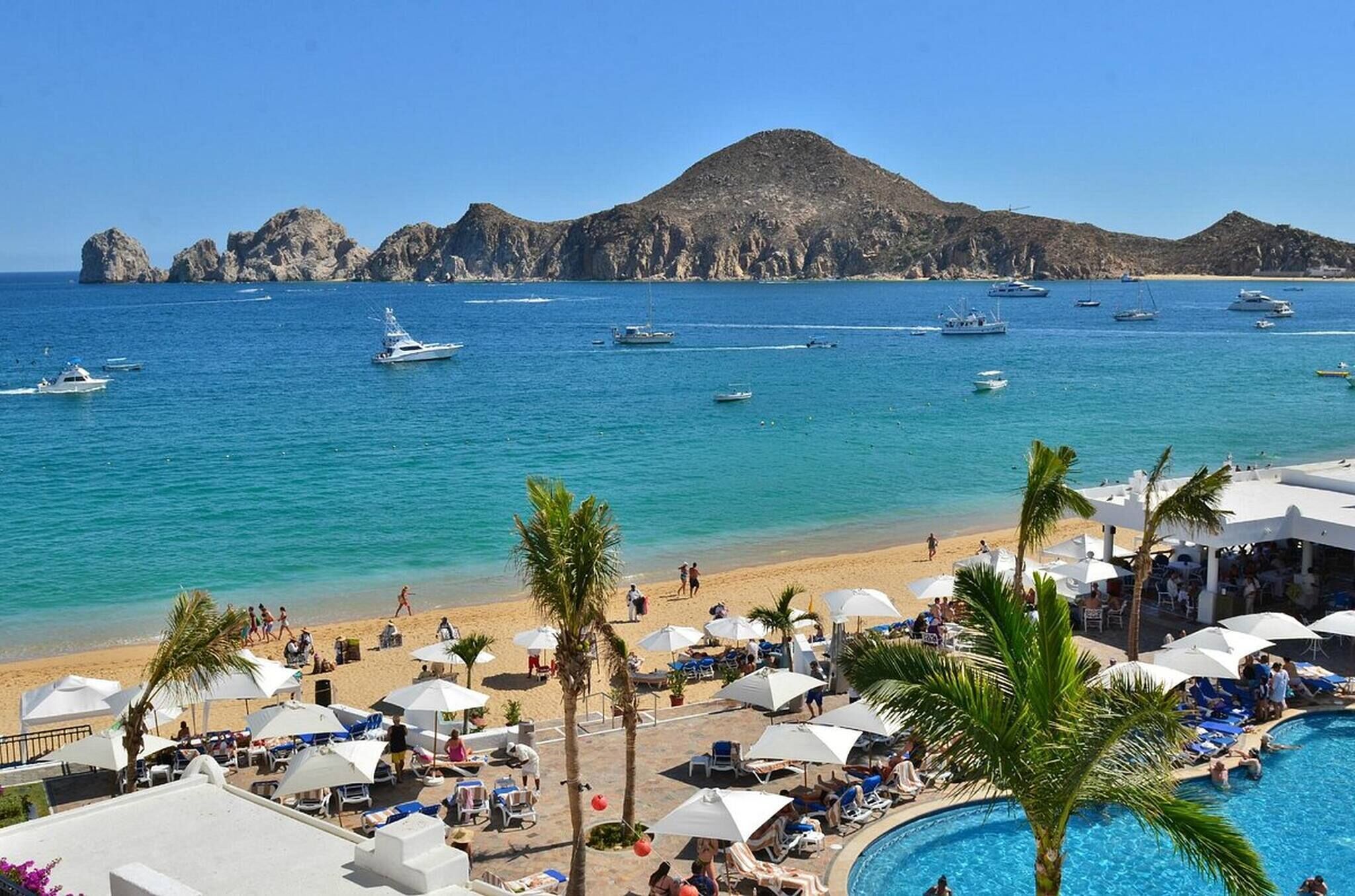 A beach resort with a pool, lounge chairs, umbrellas, and boats in the background.