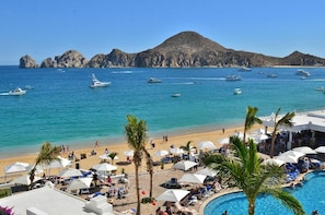 A beach resort with a pool, lounge chairs, umbrellas, and boats in the background. - Cozy Cabo Condo 2br, Shared Pool, Gated Community, 20 Walk to Beach (Cabo San Lucas)