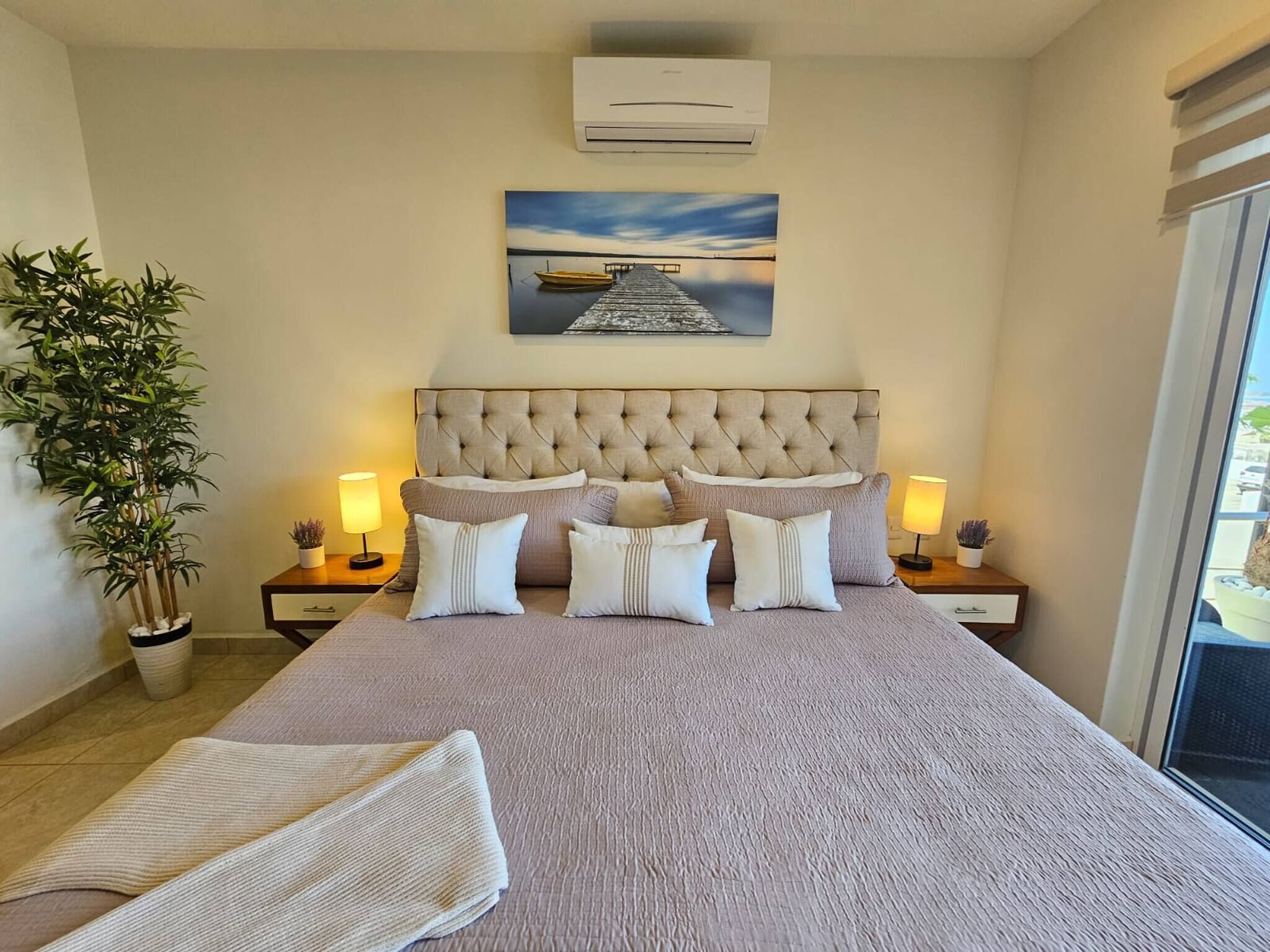 A bedroom with a large bed, two bedside tables, a potted plant, and a painting on the wall.