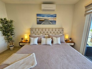 A bedroom with a large bed, two bedside tables, a potted plant, and a painting on the wall. - Cozy Cabo Condo 2br, Shared Pool, Gated Community, 20 Walk to Beach (Cabo San Lucas)
