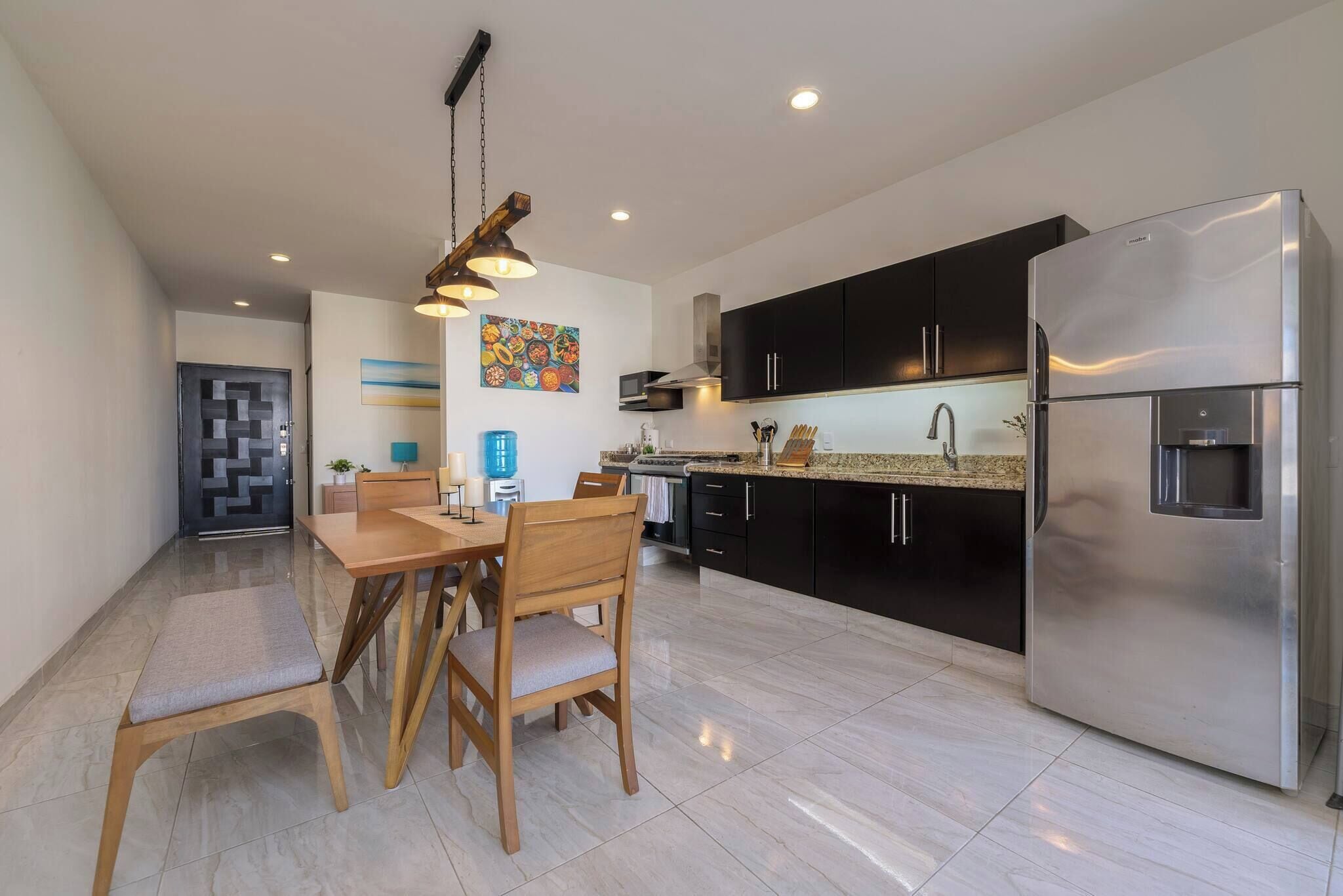 A modern kitchen with a stainless steel refrigerator, black cabinets, and a dining area with wooden chairs and a table.