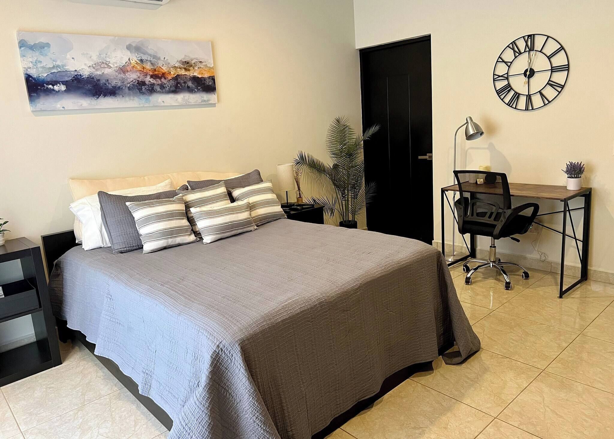 A bedroom with a bed, a desk, a chair, a clock, and a painting on the wall.