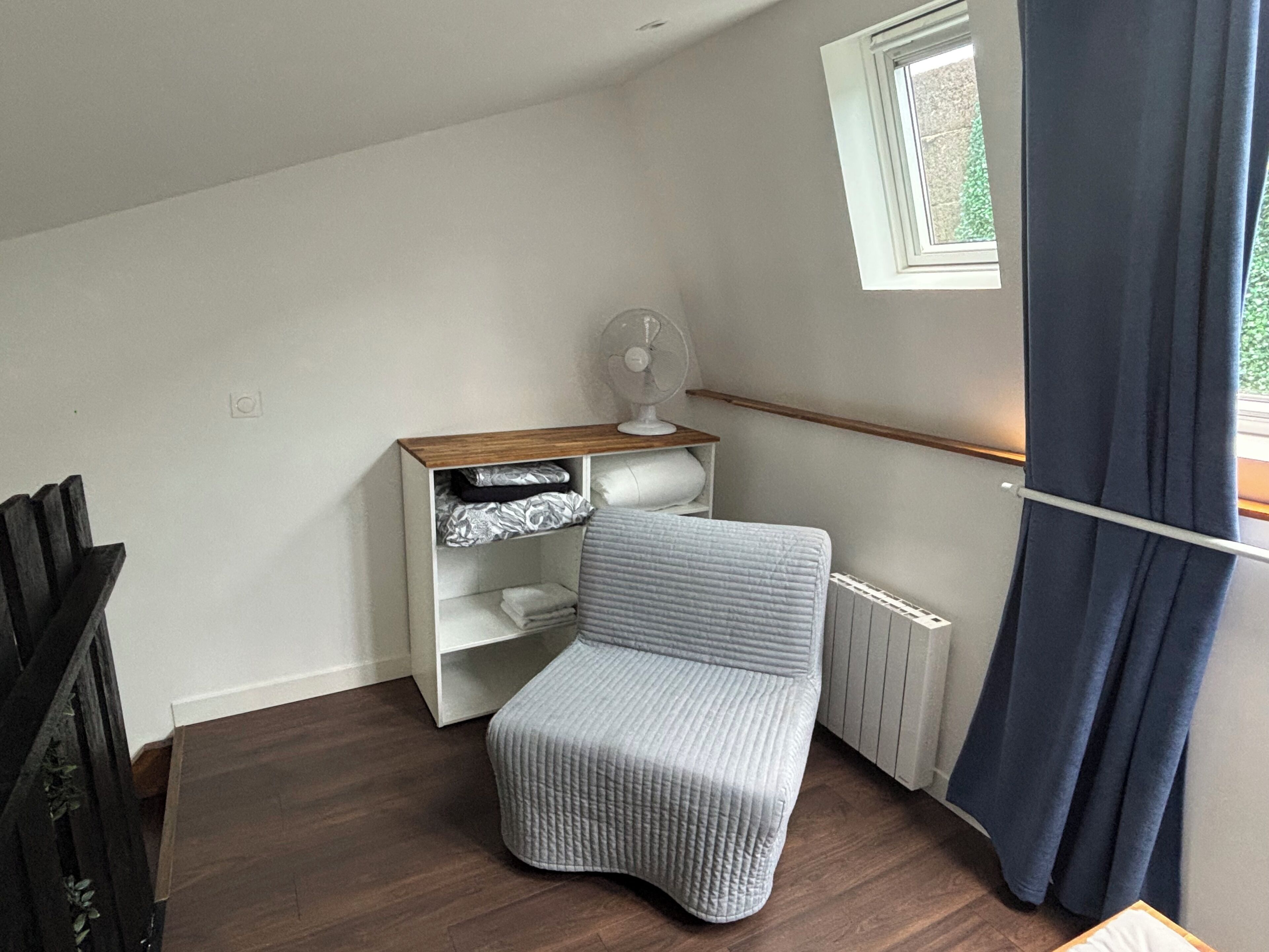 A small, cozy room with a sloped ceiling, a radiator, a white shelf unit, a grey upholstered chair, and a window with a white frame.