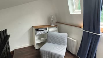 A small, cozy room with a sloped ceiling, a radiator, a white shelf unit, a grey upholstered chair, and a window with a white frame.