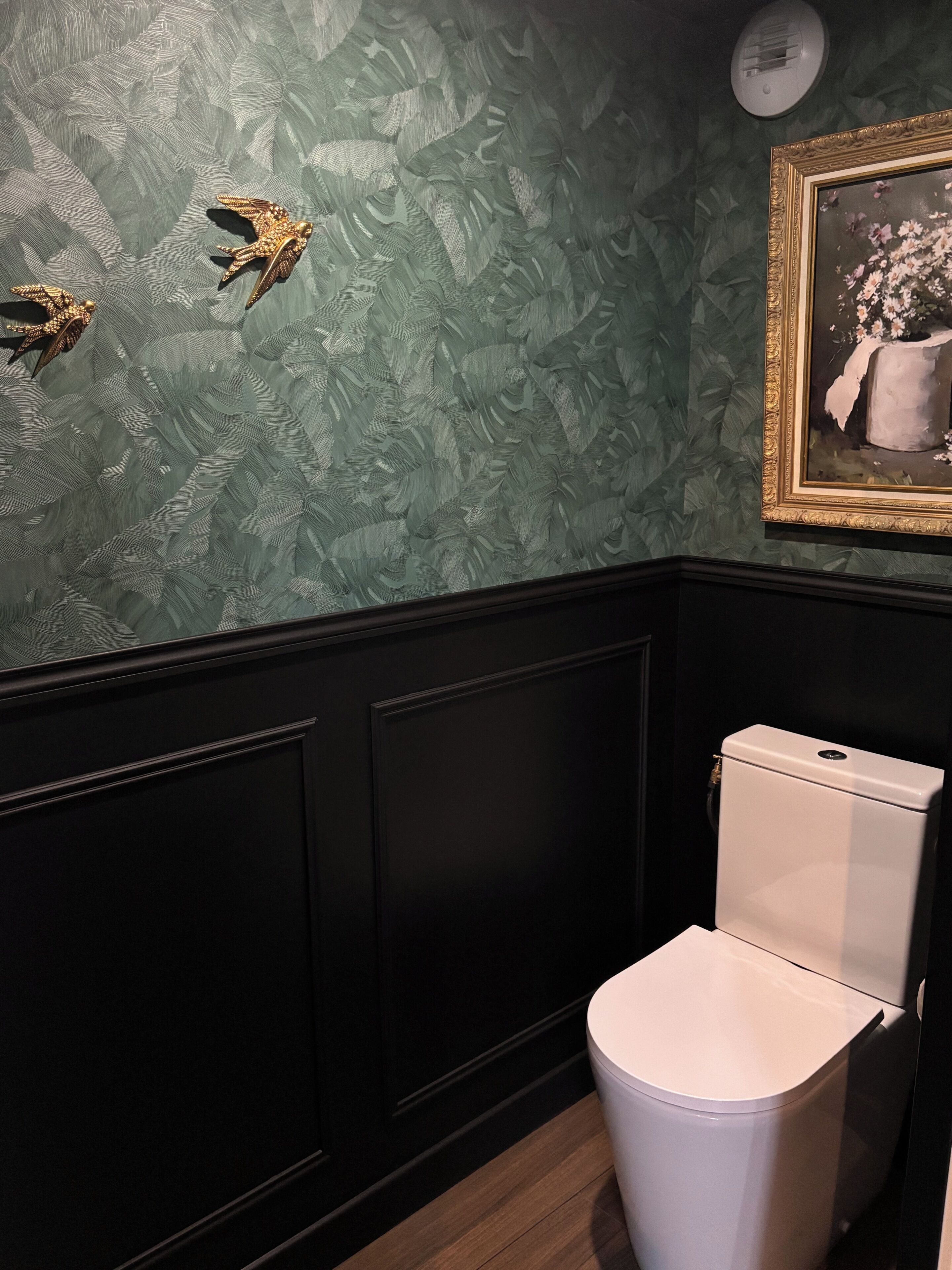 Une salle de bain avec des toilettes blanches, un plancher en bois foncé et un mur orné d’un motif de feuilles vertes.