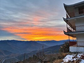 Edifício tradicional de vários andares com varanda, ao fundo montanhas ao pôr do sol.