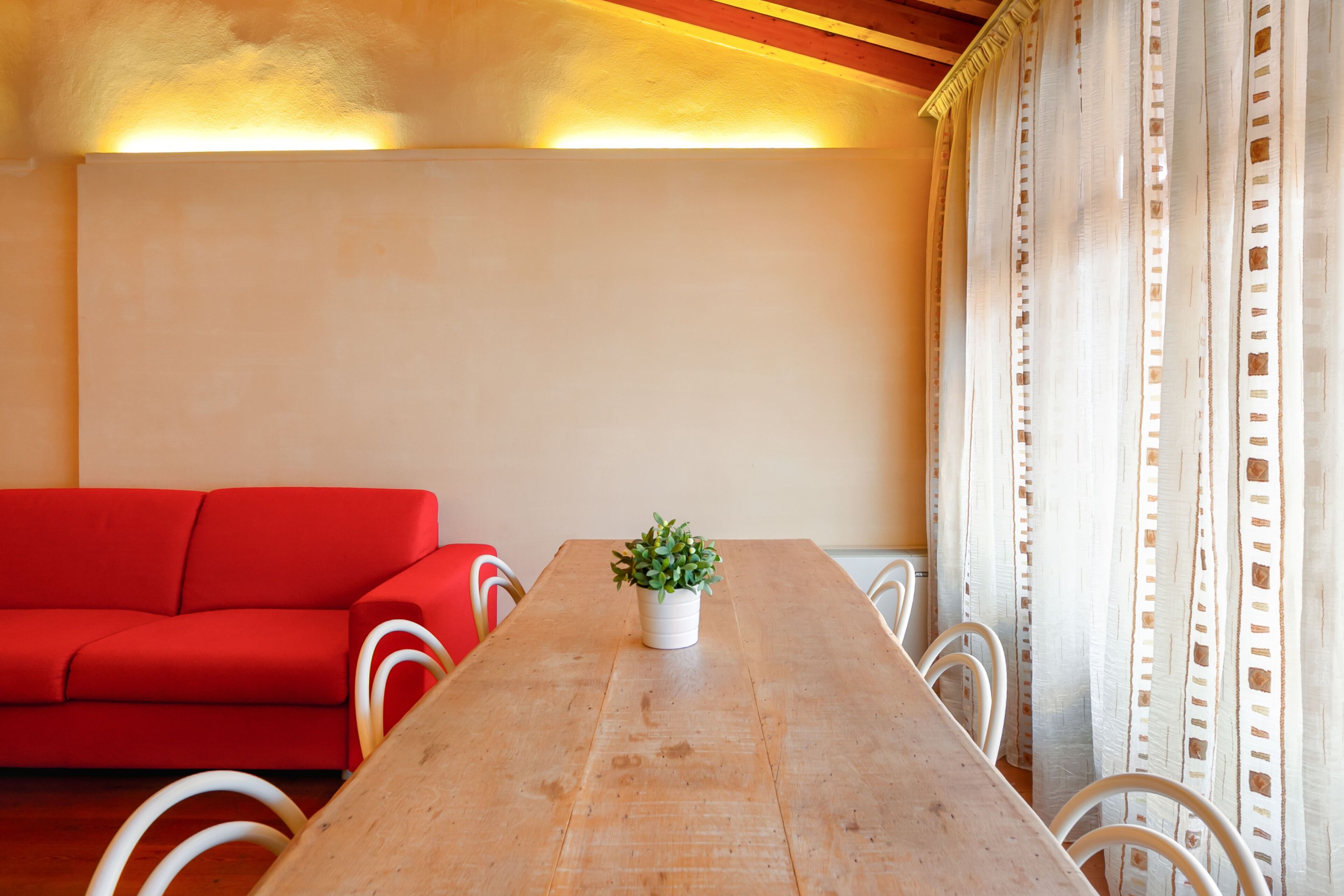 A room with a wooden dining table, a red sofa, white chairs, and a potted plant.