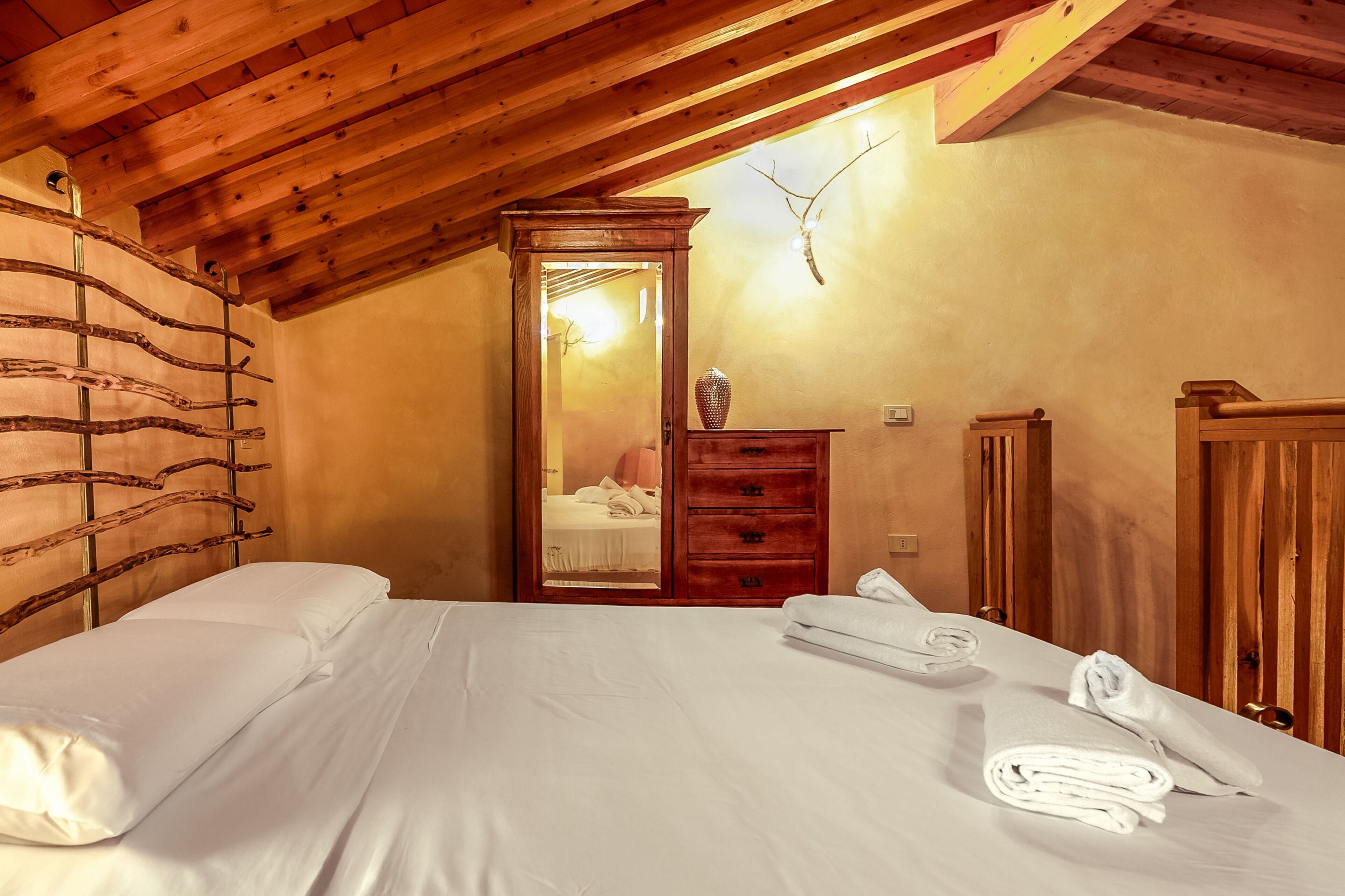A bedroom with a wooden ceiling, a bed with white linens, a wooden dresser, and a mirror reflecting a room with a bed and a lamp.