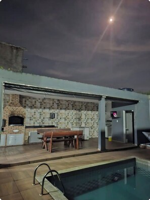 An outdoor patio with a brick oven, a table, and a pool. - Great house in Itanhaém (Itanhaém)