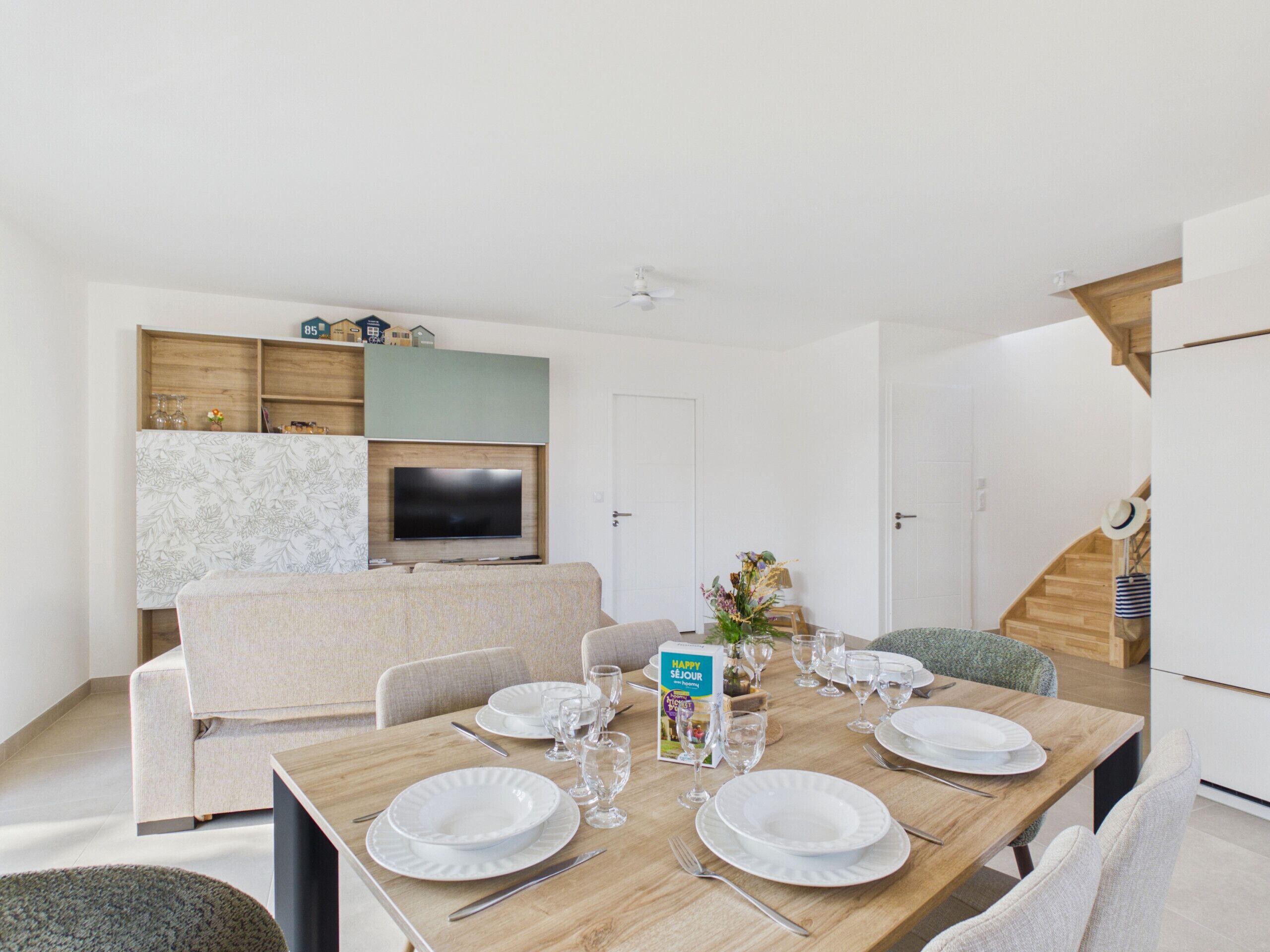 Un comedor con una mesa de madera dispuesta para cuatro personas, un sofá y un televisor montado en la pared.