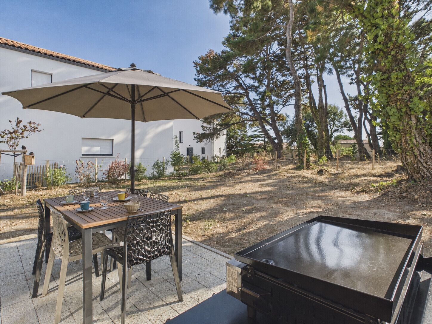 Eine Terrasse im Freien mit Tisch, Stühlen, einem Grill und einem weißen Gebäude im Hintergrund.