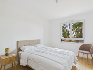 Ein Schlafzimmer mit Bett, Nachttisch, Stuhl und Fenster mit Blick auf Bäume.