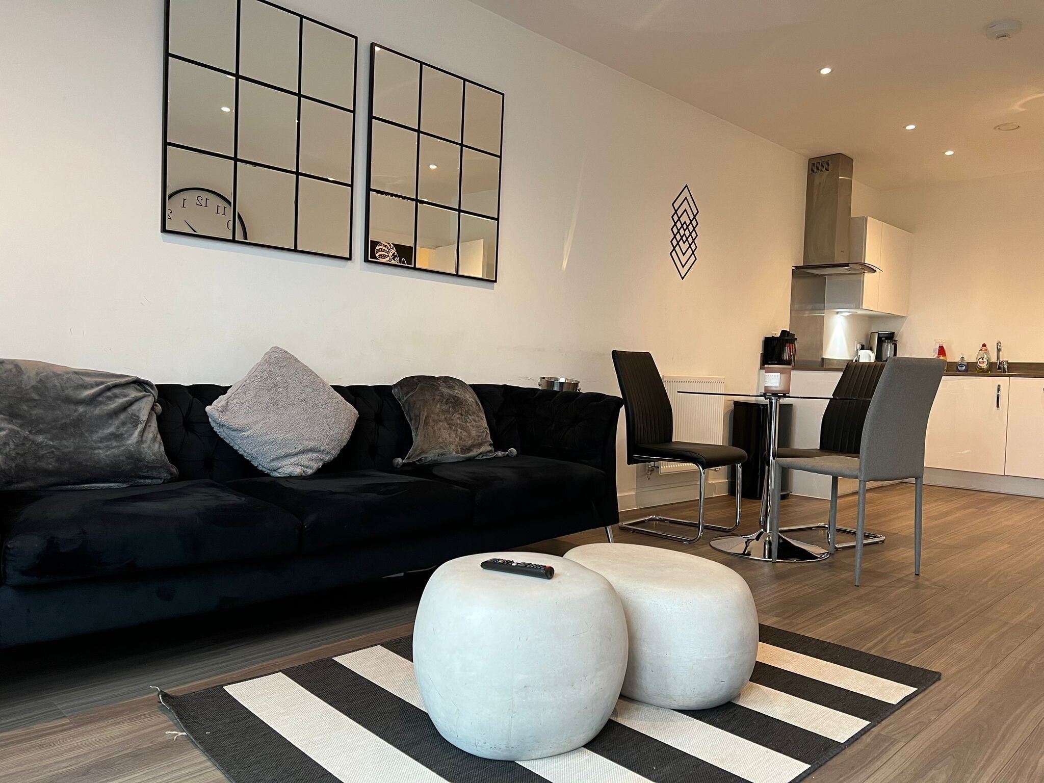Un salon moderne avec un canapé noir, deux poufs blancs, un tapis à rayures et un coin repas comprenant une table et des chaises.