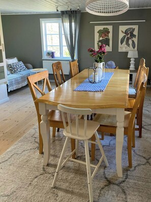A dining area with a wooden table, chairs, and a rug. There is a window with curtains, a radiator, and wall art. - Guest house on farm near Gothenburg (Västra Götalands län)