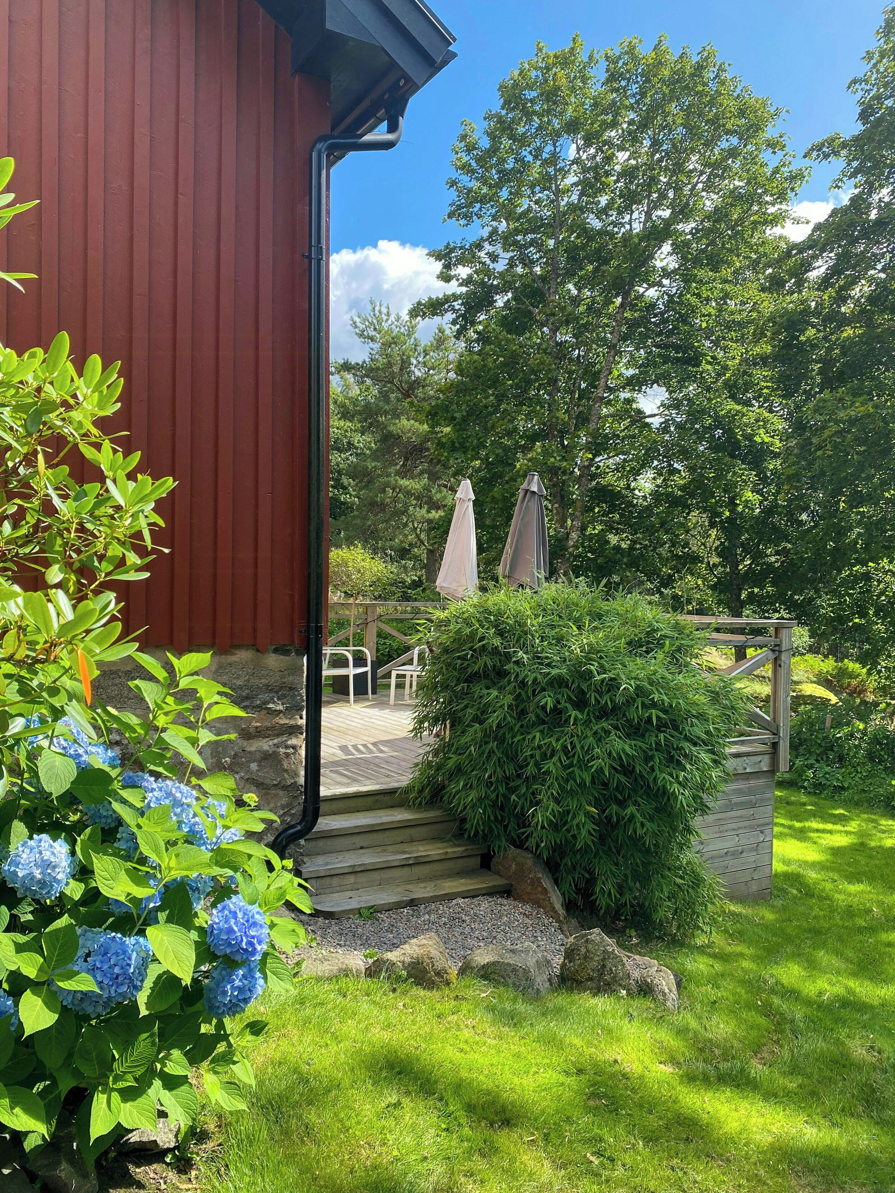 A wooden deck with outdoor furniture, a red building, and a garden with blue hydrangea flowers.
