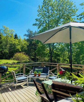 Une terrasse extérieure avec une plateforme en bois, une table entourée de chaises et un grand parasol pour faire de l’ombre.