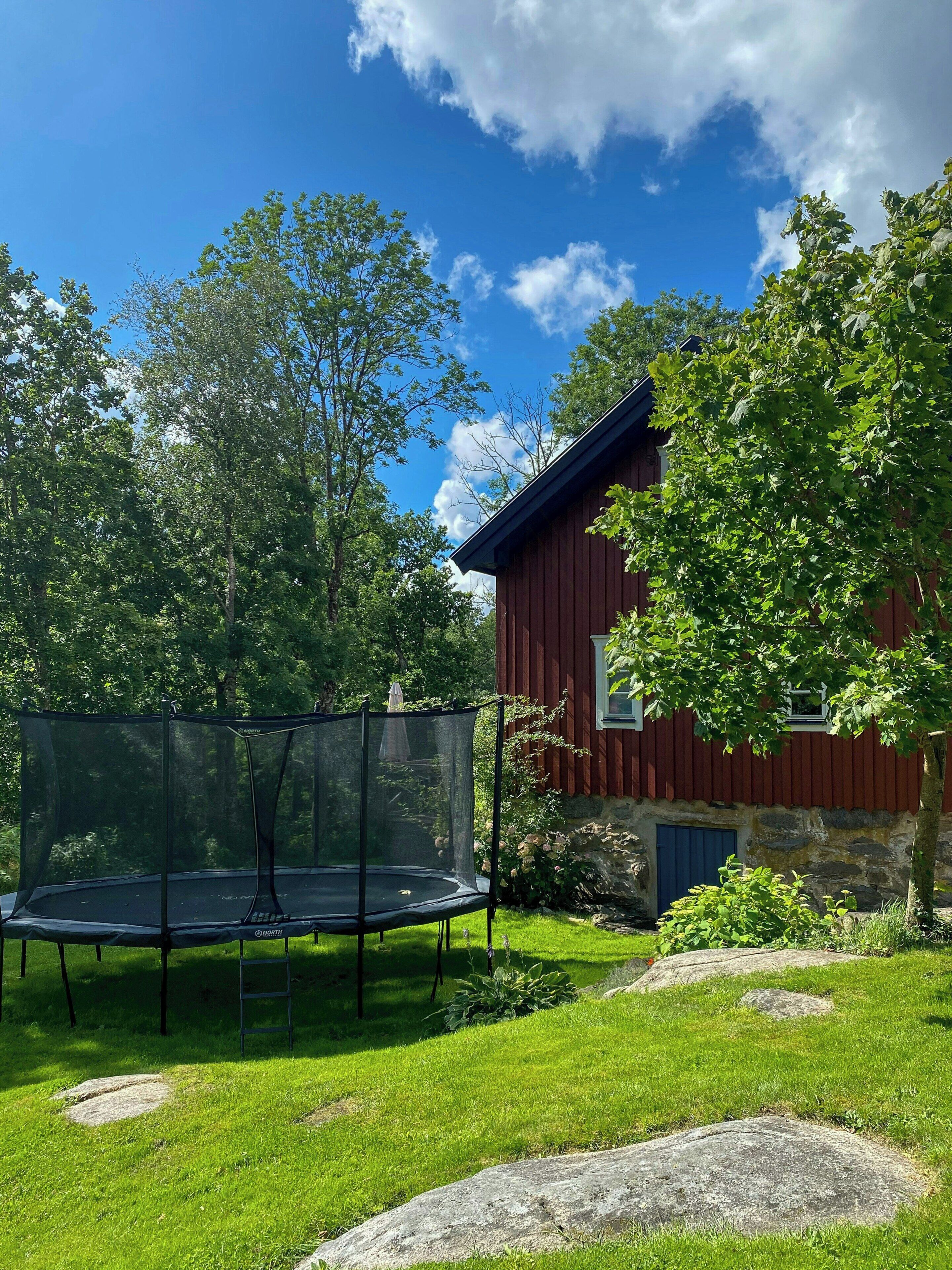 Ein rotes Holzhaus mit Steinuntergrund, umgeben von einem grünen Rasen mit Trampolin und Baum.