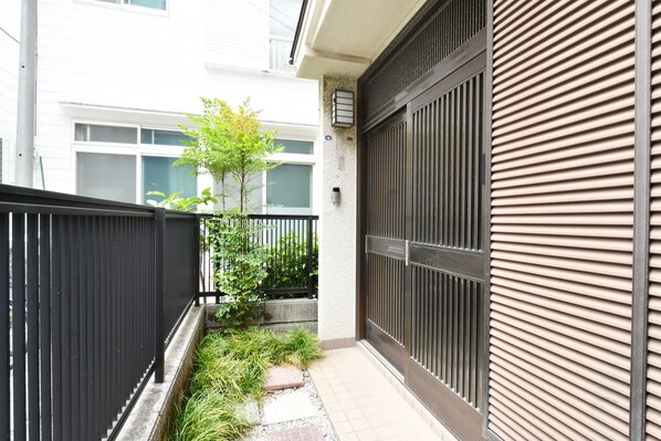 A narrow outdoor corridor with a black metal fence on one side and a beige wall with horizontal slats on the other. There is a small patch of greenery and a tree in the center.