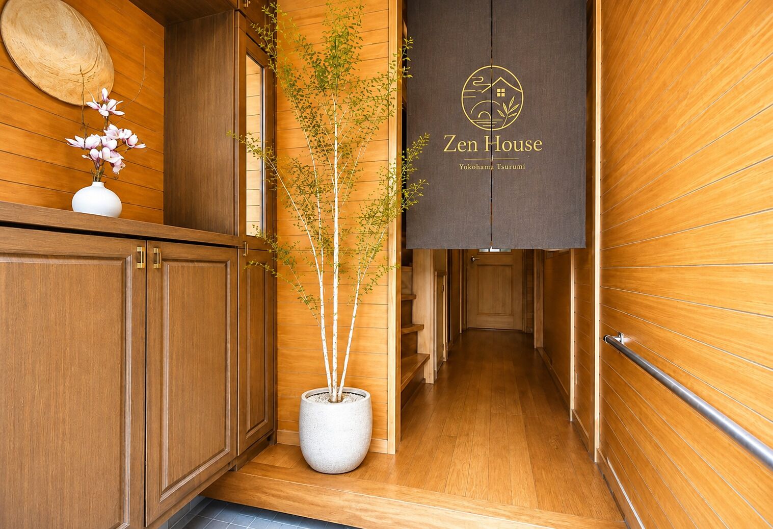 A wooden interior hallway with a potted tree, a wall-mounted sign reading 'Zen House Yokohama Tsuruumi,' and a wooden door at the end.