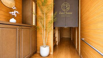 A wooden interior hallway with a potted tree, a wall-mounted sign reading 'Zen House Yokohama Tsuruumi,' and a wooden door at the end.