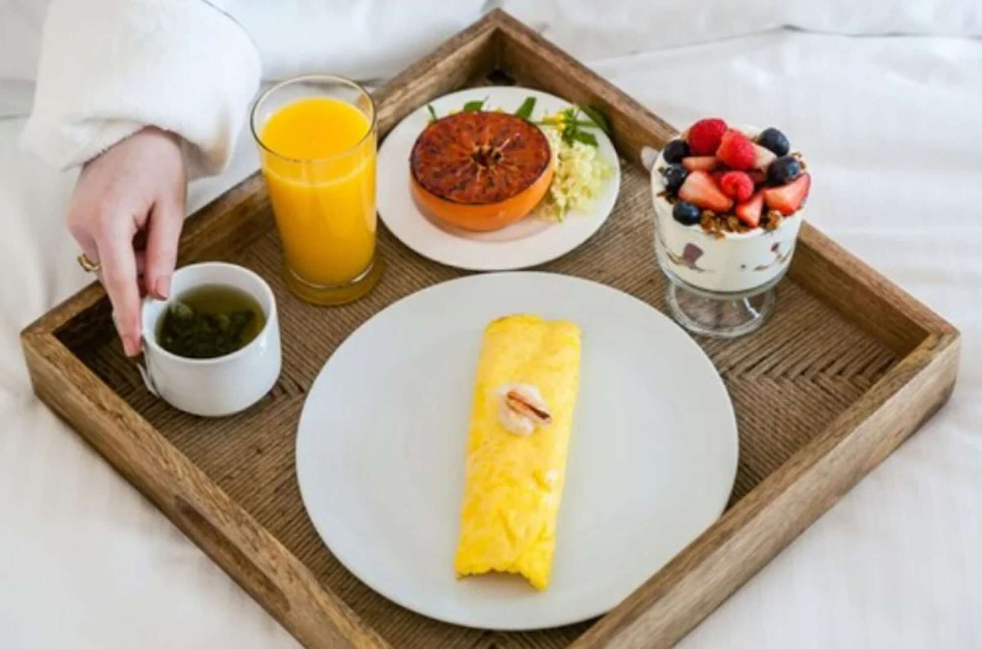 Uma bandeja de café da manhã com um omelete enrolado, um copo de suco de laranja, um prato com toranja e folhas verdes, uma xícara de chá e um copo de iogurte coberto com frutas vermelhas.