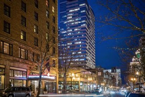 A city street at night with buildings, including a tall modern building with lit windows, and a traditional brick building with a theater. - Downtown Views | Pet-friendly & In-Room Dining (Portland)