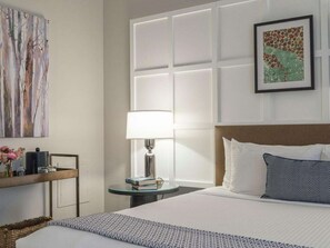 Habitación de hotel con una cama grande, una mesita de noche con lámpara, un cuadro enmarcado en la pared y un adorno sobre la cama.