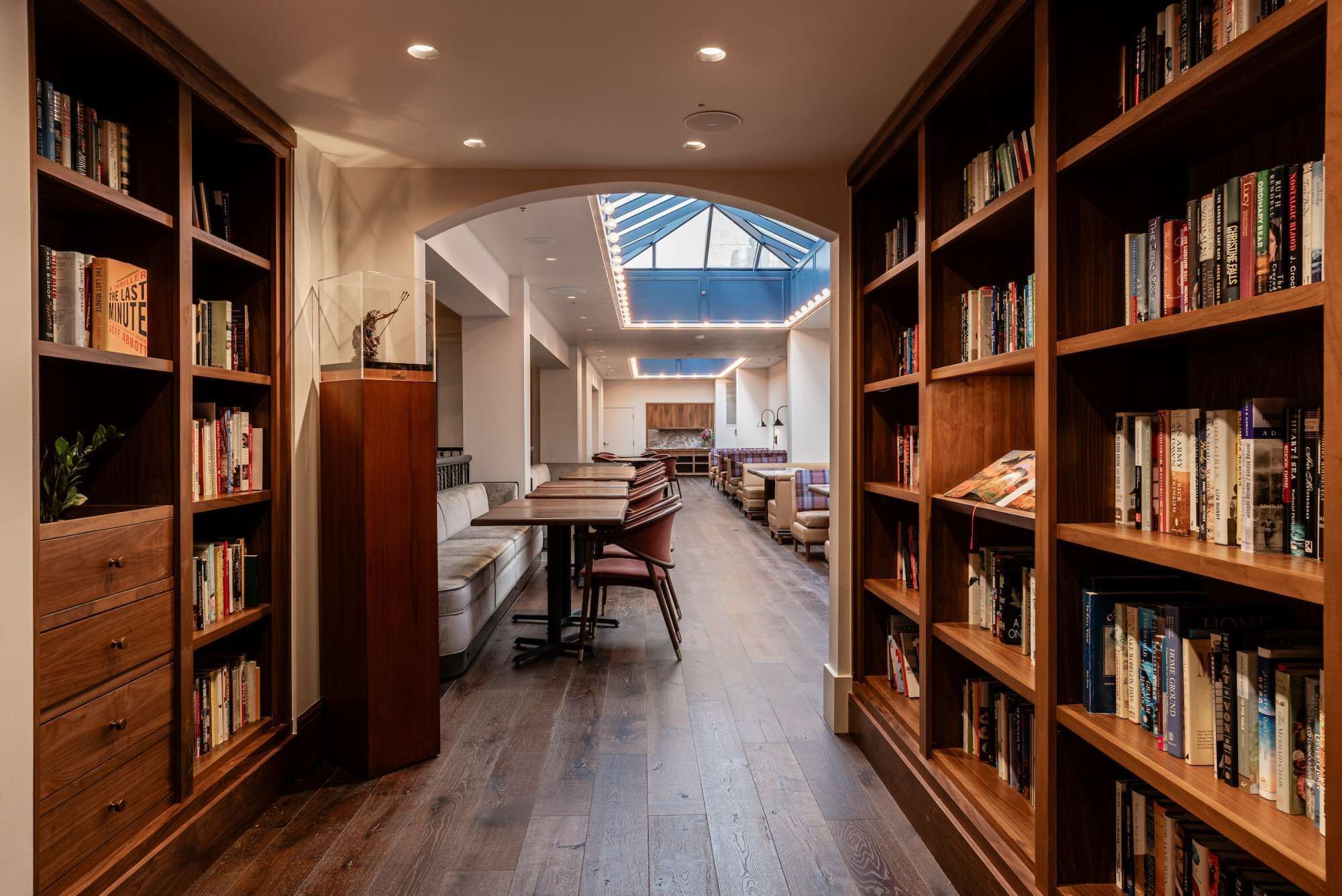 Una biblioteca con scaffali su entrambi i lati, un'area centrale con tavoli e sedie, e un soffitto con lucernario.