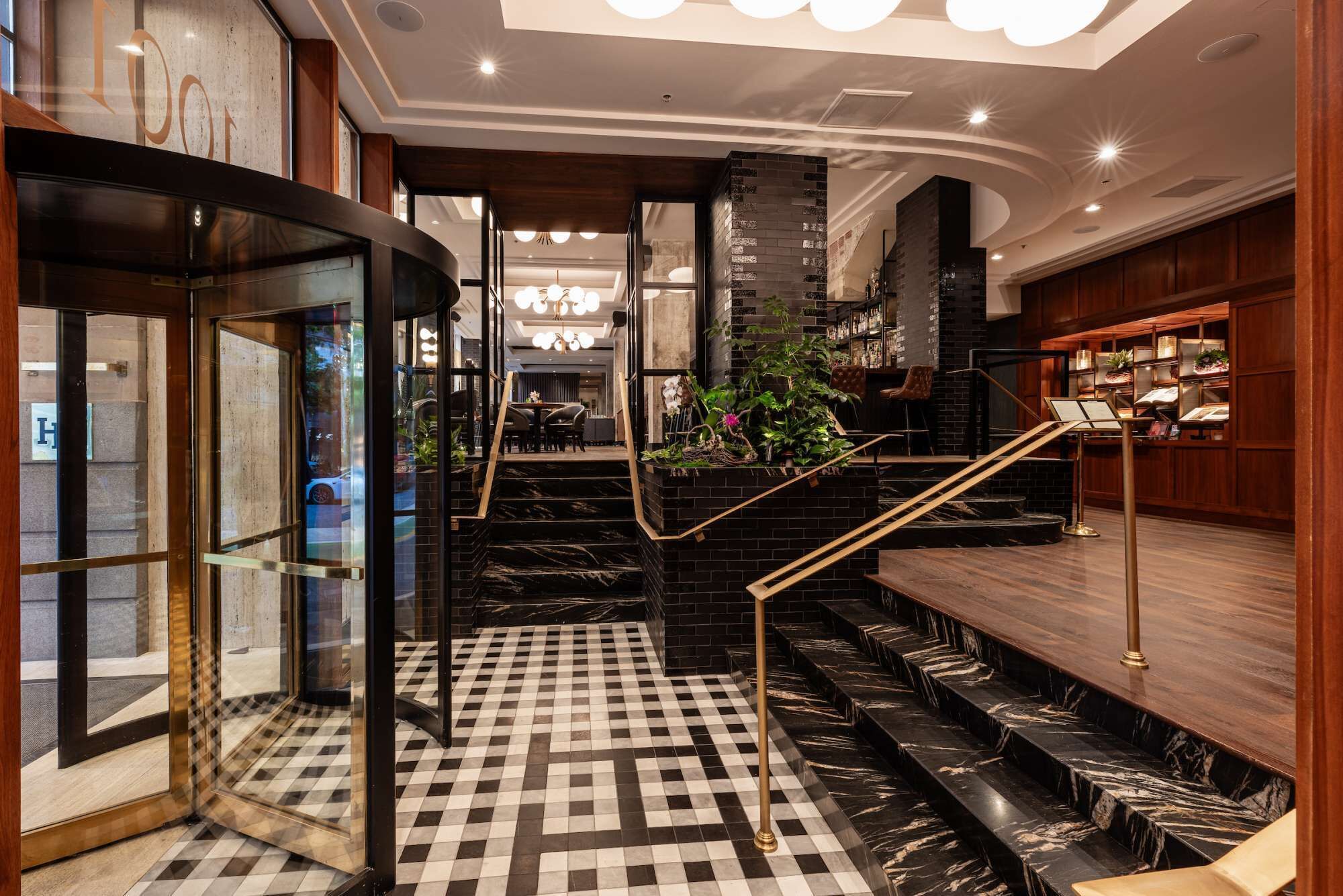 Un'accogliente hall d'albergo con pavimento a scacchi, una scala con ringhiera nera e dorata e un'area bar con mobiletti in legno.