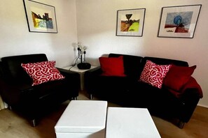Een woonkamer met twee zwarte banken, elk voorzien van een kussen met een rood-wit patroon, een witte salontafel en drie kunstwerken in lijsten aan de muur.