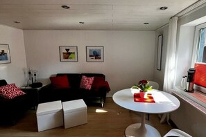 Una sala de estar con un sofá negro, un reposapiés blanco y una mesita pequeña con flores. Hay cuadros en la pared y una ventana con cortinas.