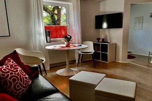 Ein modernes Wohnzimmer mit einer Couch, einem Esstisch mit Stühlen, einem flachbildfernseher an der Wand und einem Fenster mit Jalousien.