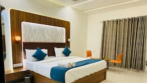 Ein modernes Hotelzimmer mit einem Holz-Kopfteil, einem großen Bett mit weißer und blauer Bettwäsche, zwei orangefarbenen Sesseln und einem Vorhang.