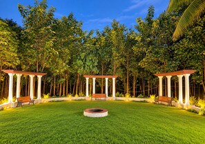 A well-maintained lawn with three wooden benches and a circular fire pit, surrounded by trees and a clear sky.