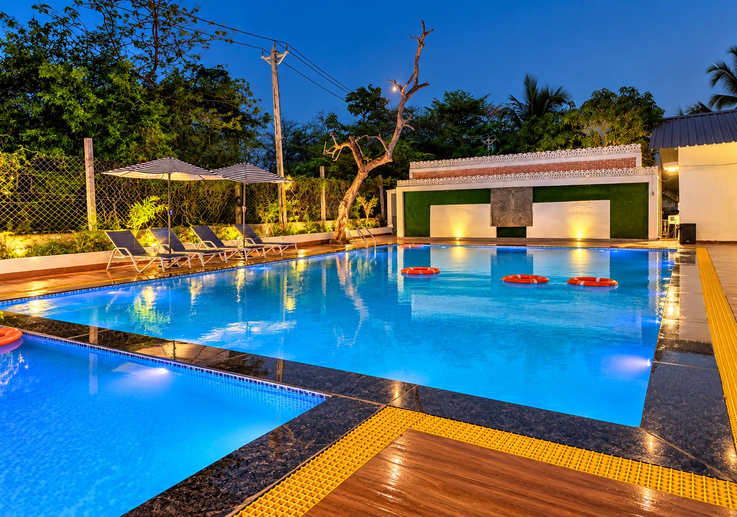 Una piscina con sillas de descanso y un flotador salvavidas, rodeada de vegetación y un edificio con pared verde.