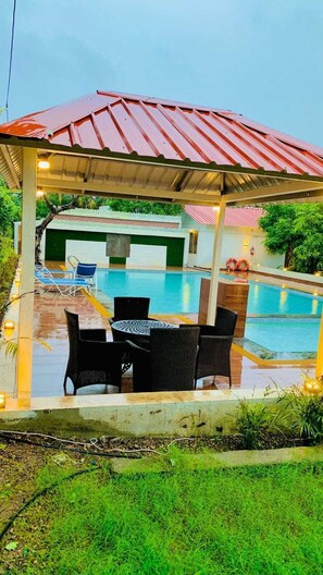 Un espace extérieur aménagé avec des sièges, protégé du soleil, et une piscine en arrière-plan.
