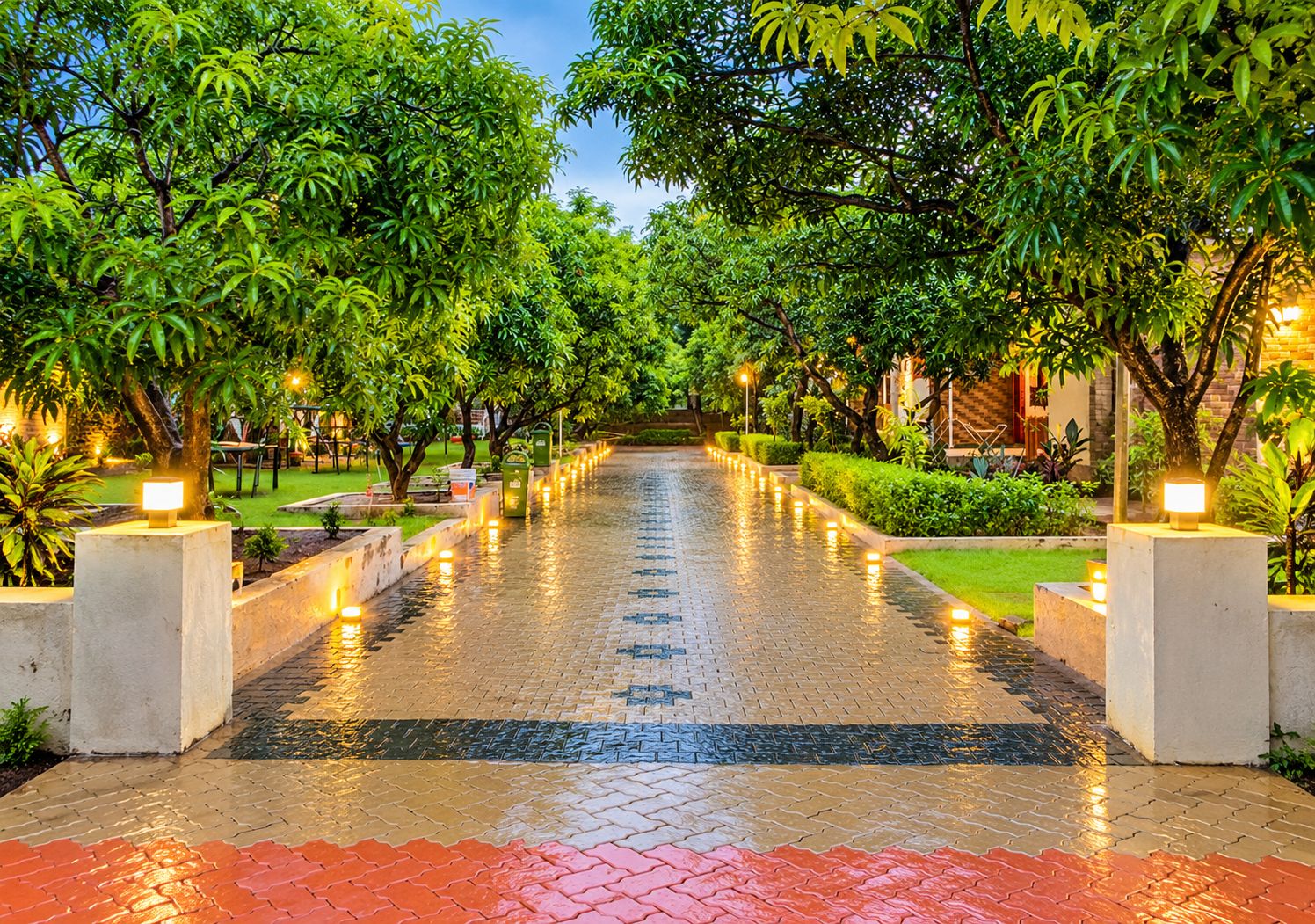Un chemin pavé, bordé d’arbres et éclairé par des luminaires au sol, mène à l’entrée d’un bâtiment.