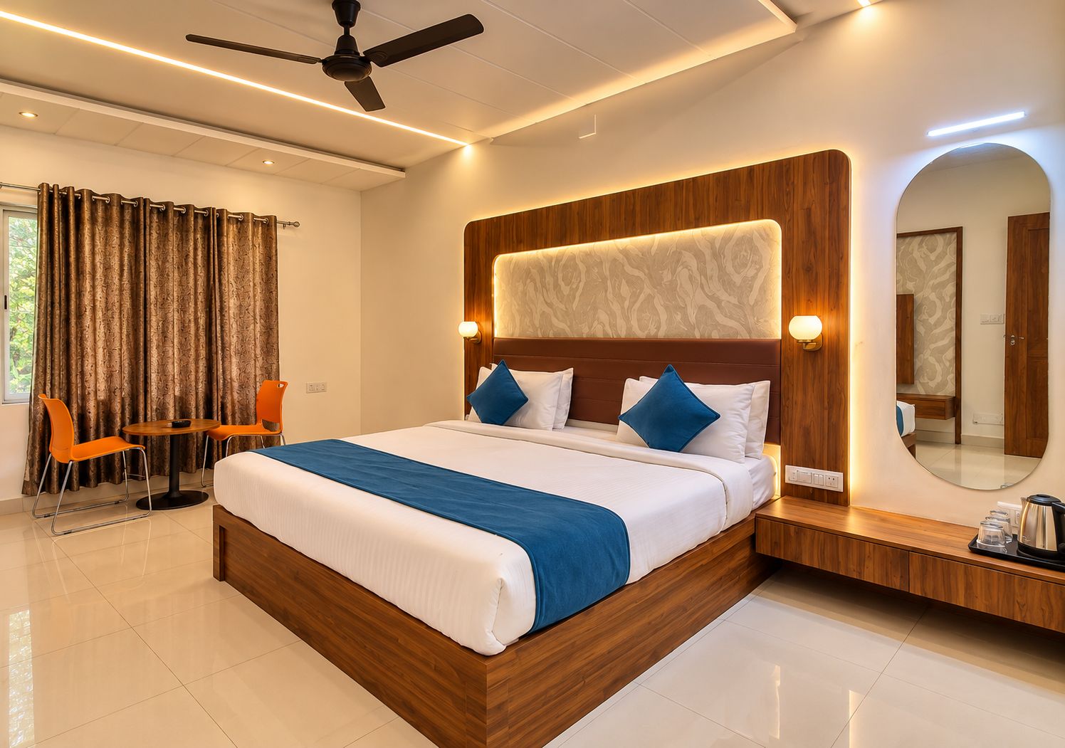 Una habitación de hotel moderna con una cama grande, un ventilador de techo, una zona de estar con sillas y un espejo amplio.