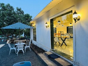An outdoor seating area with tables and chairs, a striped umbrella, and a glass door leading to an interior space with tables and chairs. - Sorathgadh Resort Sasan Gir (Sasan Gir)