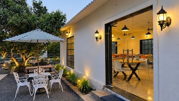 An outdoor dining area with tables and chairs, a striped umbrella, and decorative lighting.
