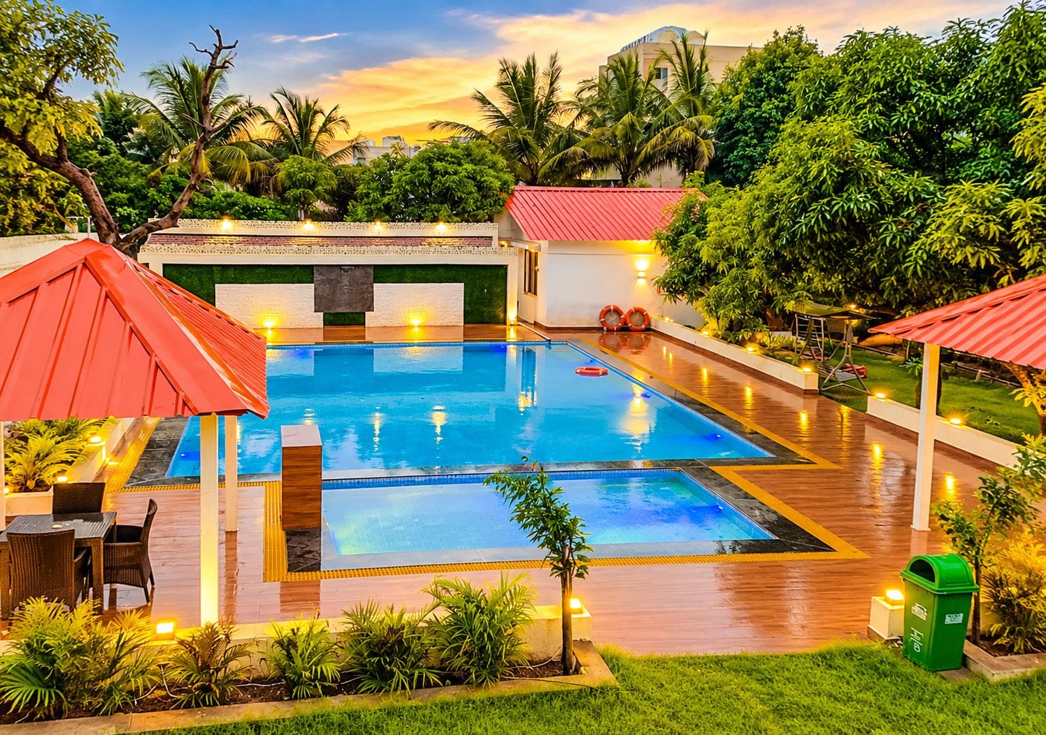 Une piscine rectangulaire dotée d’un espace abrité avec un toit rouge, entourée de verdure et bordée d’un bâtiment blanc également surmonté d’un toit rouge.