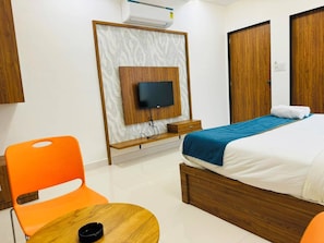 Een hotelkamer met een bed, een aan een houten paneel bevestigde televisie, een kleine tafel en een oranje stoel.