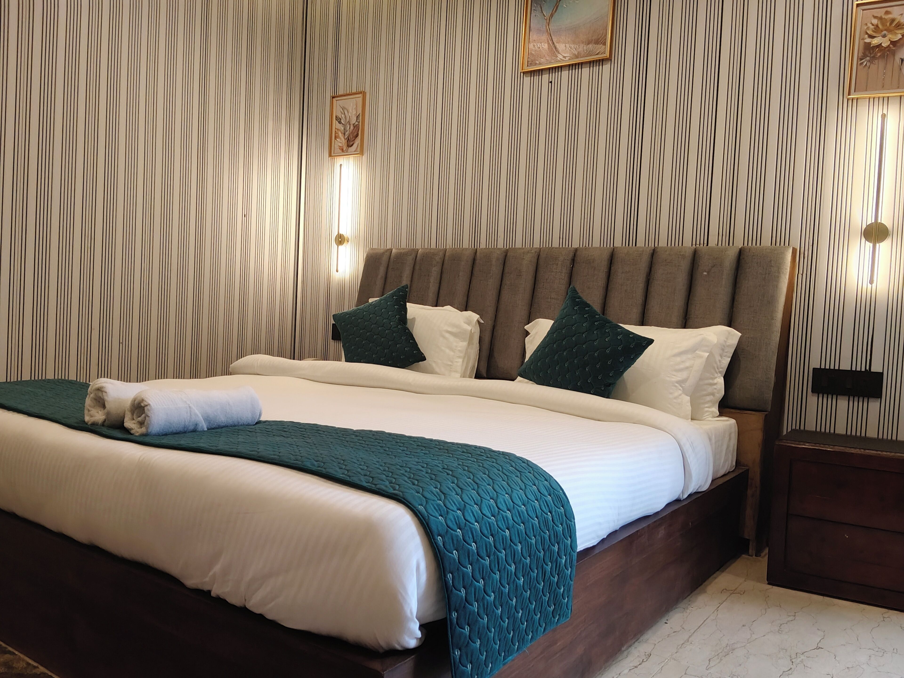 A neatly made bed with a green blanket and a rolled towel, a headboard with multiple pillows, and a wall with vertical lines and two wall-mounted lights.