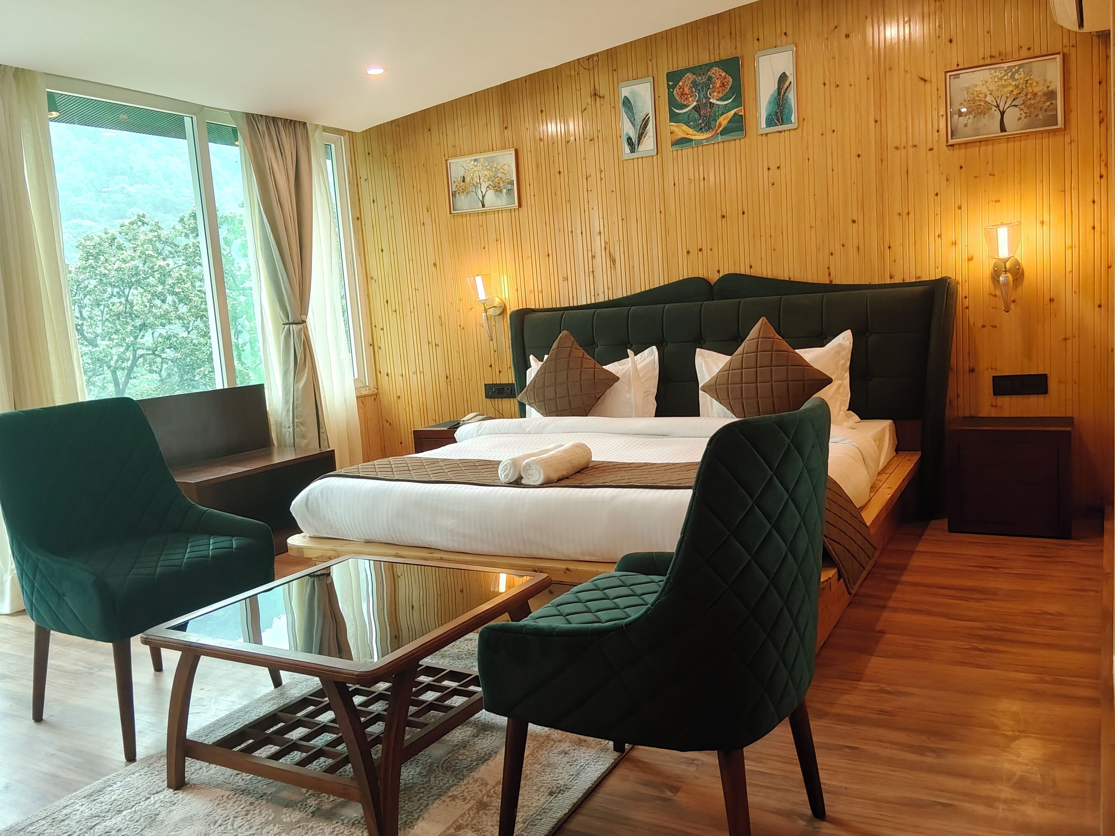 A hotel room with a large bed, two green armchairs, a glass-top table, wooden flooring, and a wooden paneled wall with artwork.